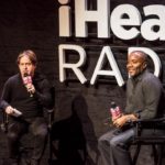 Two speakers seated in conversation on an iHeart Radio stage
