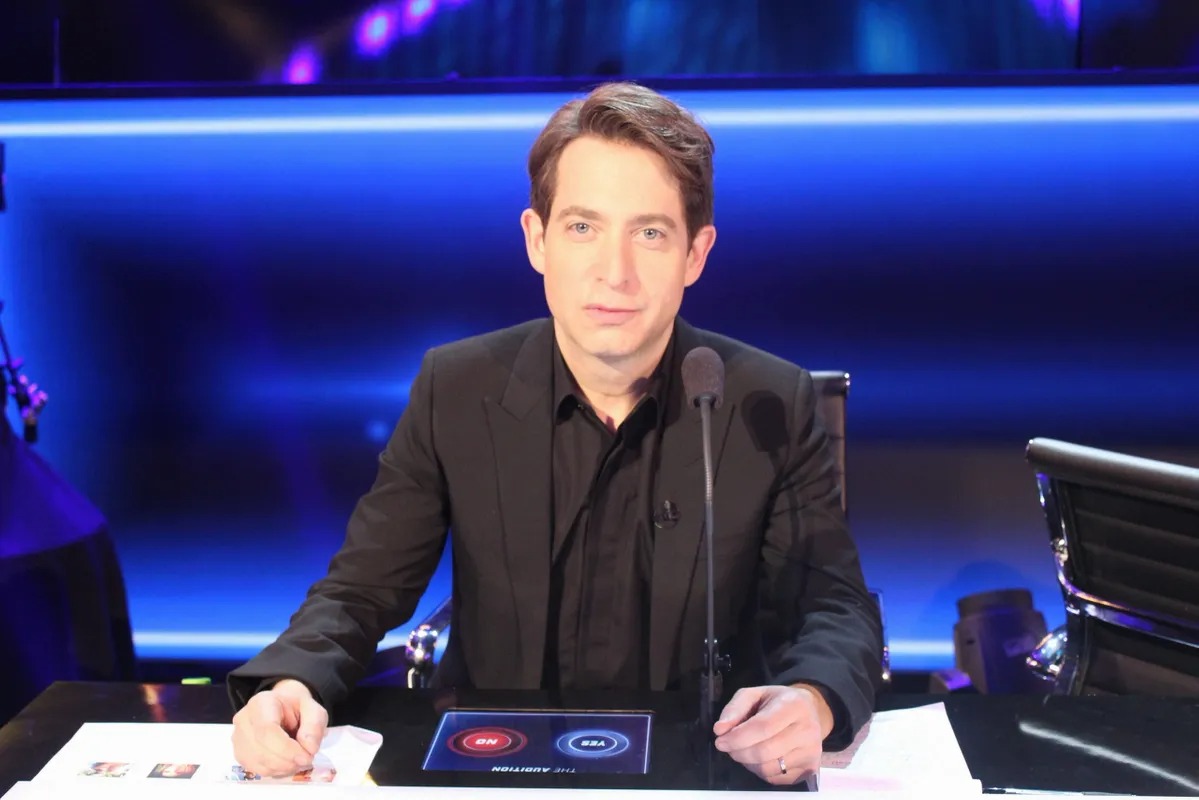 Man in a black suit seated at a judges desk with a tablet