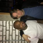 Two men posing together beside a brick wall