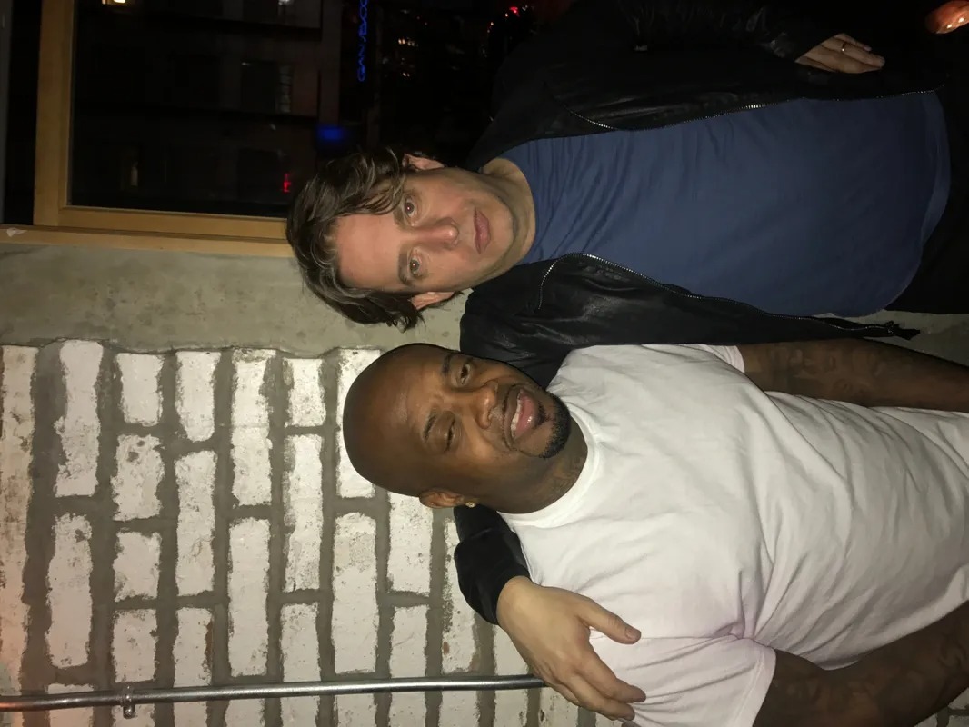Two men posing together beside a brick wall