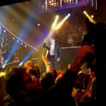 Performer singing on stage with crowd silhouettes in front